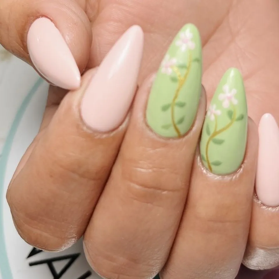Uñas almendradas en rosa pastel con dos uñas verdes y ramas con flores pintadas a mano.